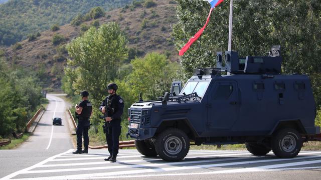 Am Rande einer asphaltierten Strasse in den Bergen steht ein kosovarischer Polizeibeamter und ein Soldat der NATO-Friedensgruppe vor einem Jeep und überwachen die Strasse.
