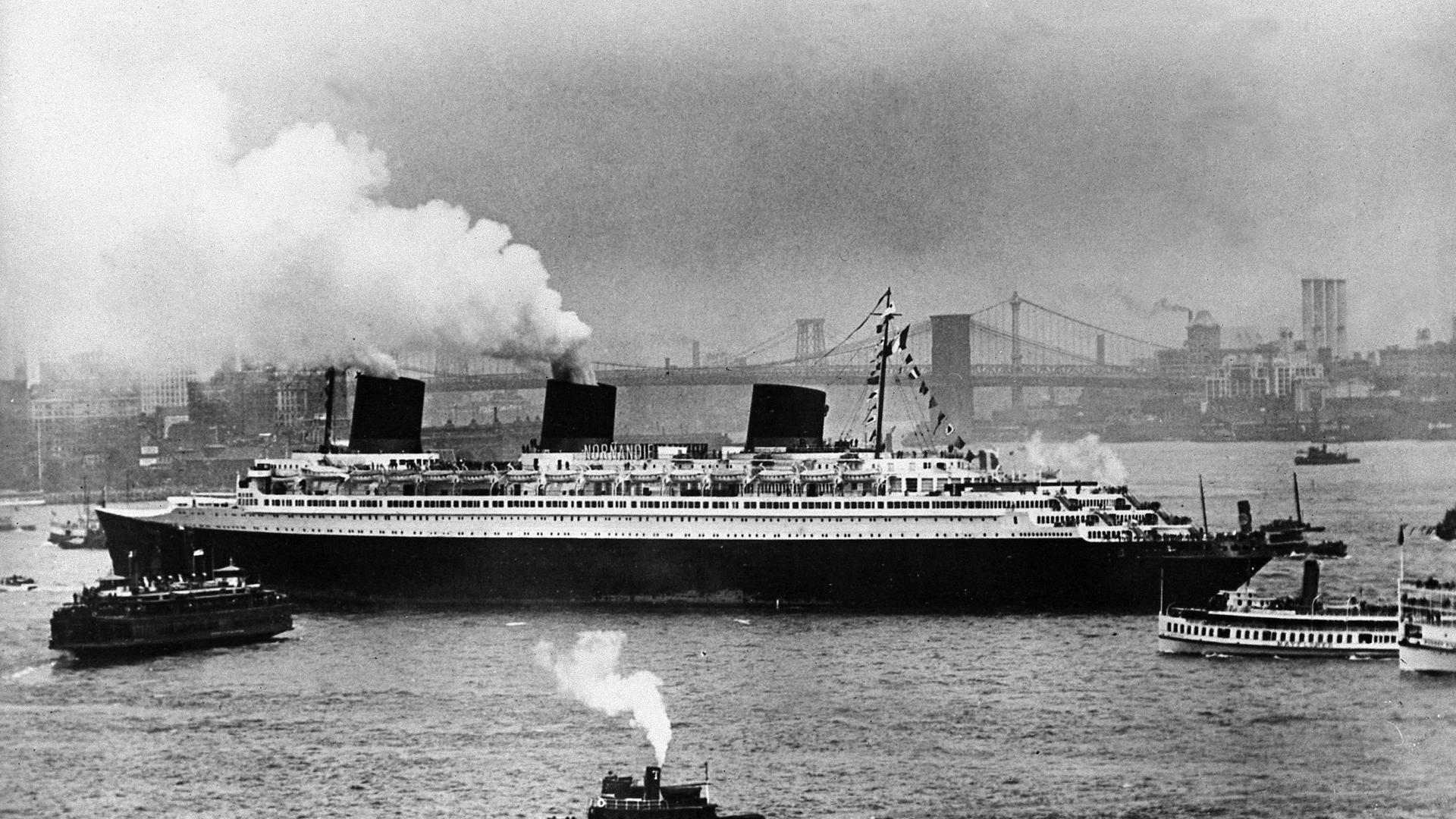 Der französische Luxusliner "Normandie" bei seiner Ankunft im Hafen von New York im August 1939. Aus zwei von drei Schornsteinen kommt Rauch. Mehrere kleinere Schiffe fahren um die "Normandie" herum. Im Hintergrund ist eine Brücke und die Silhouetten von Gebäuden zu sehen.