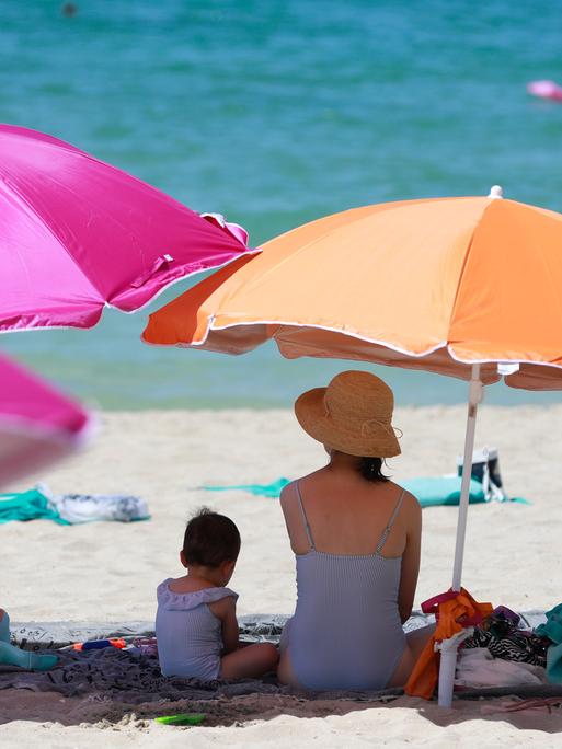 Eine Frau sitzt neben einem Kind unter einem Sonnenschirm an einem warmen Sommertag am Strand.