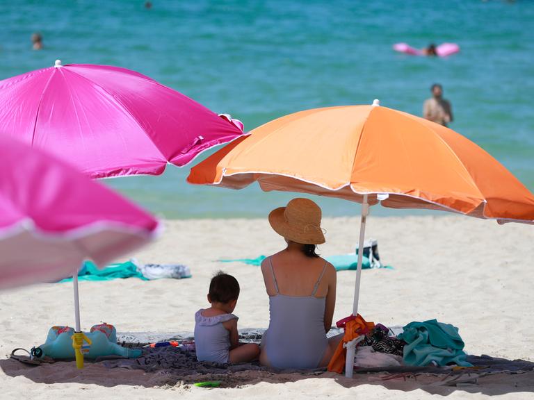 Eine Frau sitzt neben einem Kind unter einem Sonnenschirm an einem warmen Sommertag am Strand.