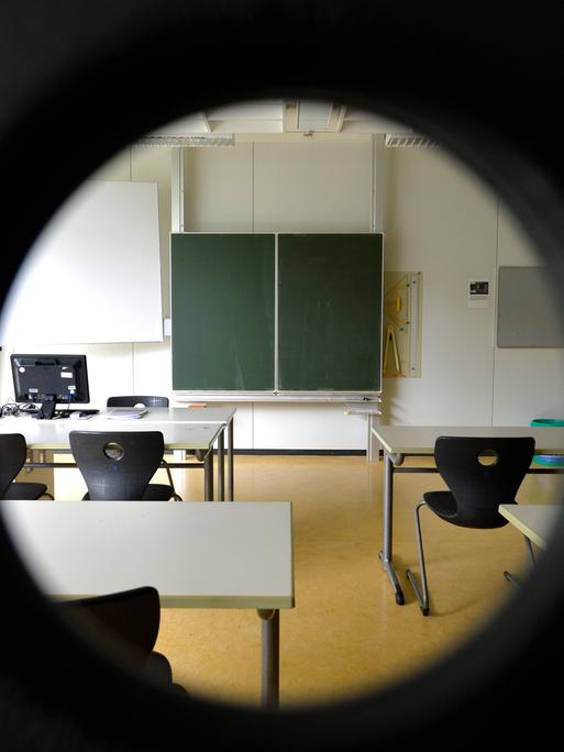 Blick durch ein rundes Loch in ein Klassenzimmer mit hochgestellten Stühlen und ohne Schüler