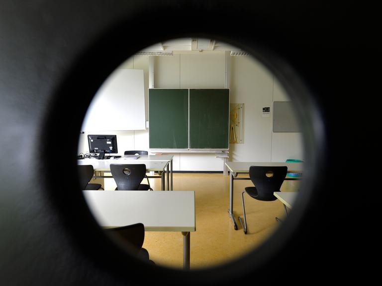 Blick durch ein rundes Loch in ein Klassenzimmer mit hochgestellten Stühlen und ohne Schüler