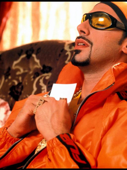 Sacha Baron Cohen als Rapper, auffällige Brille, Bling-Bling, Goldkette, Porträt, an sein Herz gedrückt; alternativer Titel.