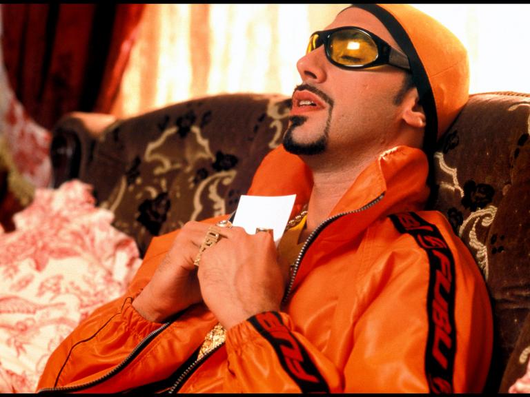 Sacha Baron Cohen als Rapper, auffällige Brille, Bling-Bling, Goldkette, Porträt, an sein Herz gedrückt; alternativer Titel.