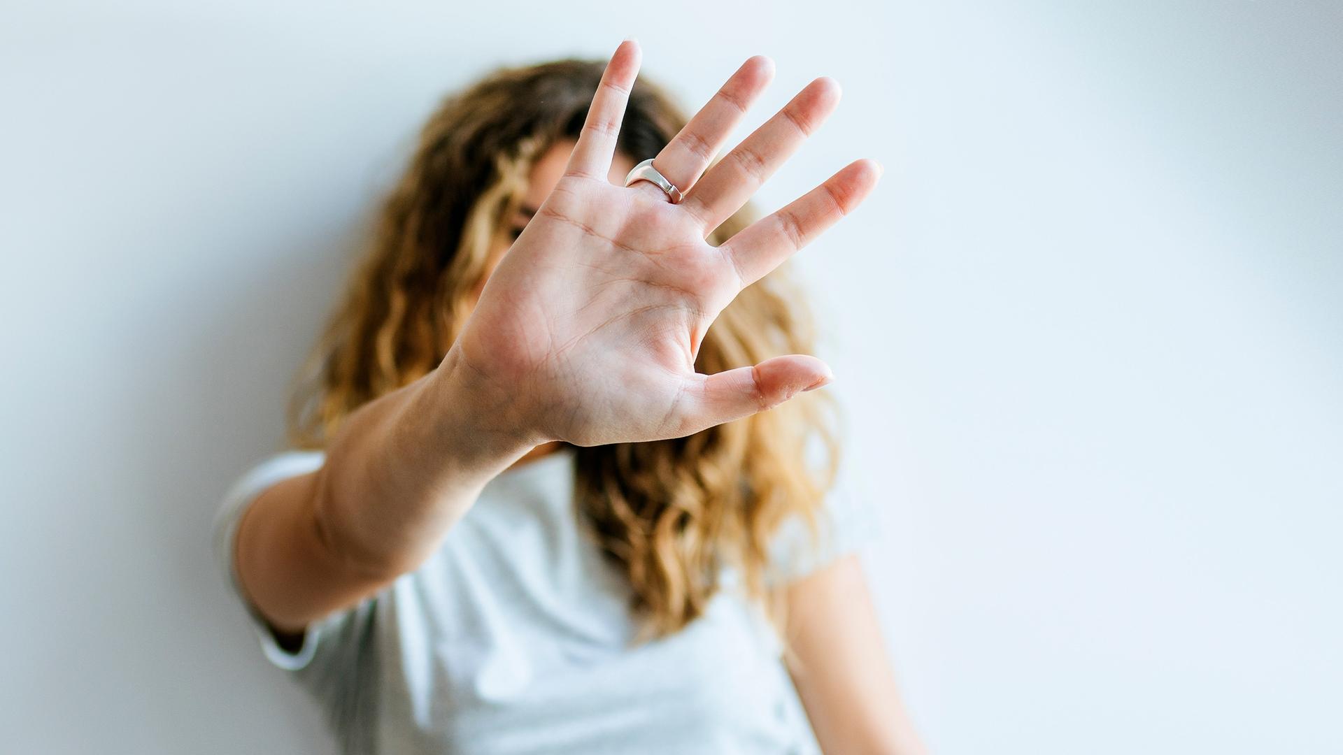 Porträt einer Frau, die ihre Hand abwehrend in die Kamera hält und dabei ihr Gesicht verdeckt
