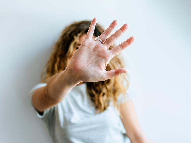 Porträt einer Frau, die ihre Hand abwehrend in die Kamera hält und dabei ihr Gesicht verdeckt
