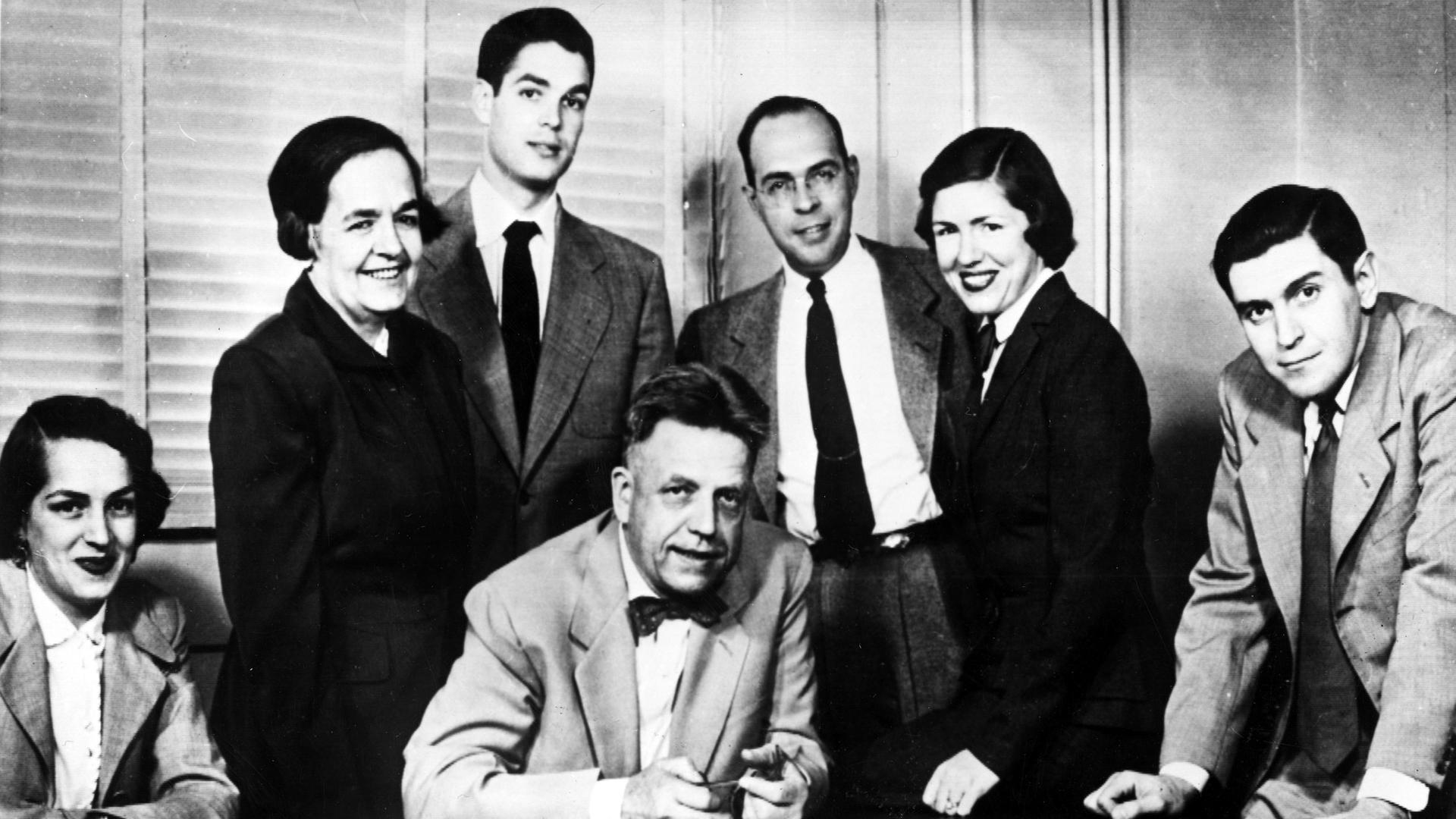Das Schwarzweißfoto zeigt einen Mann am Schreibtisch umgeben von je drei Frauen und Männern.