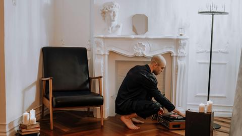 Ein junger Mann mit kurzen Haaren und dunkler, bequemer Kleidung sitzt vor dem Kamin und bedient seinen Plattenspieler.