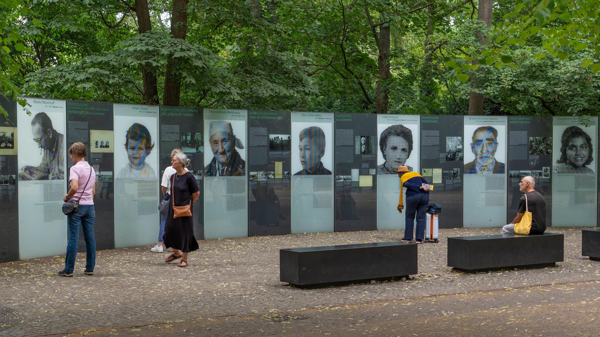 Mehrere Menschen stehen vor einem Denkmal, das an die von Nationalsozialisten ermordeten Sinti und Roma in Europa erinnert. Auf Gedenktafeln sind die Gesichter mehrerer Opfer und deren Geschichten abgebildet. 