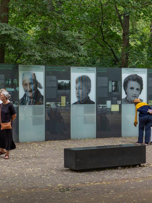 Mehrere Menschen stehen vor einem Denkmal, das an die von Nationalsozialisten ermordeten Sinti und Roma in Europa erinnert. Auf Gedenktafeln sind die Gesichter mehrerer Opfer und deren Geschichten abgebildet. 