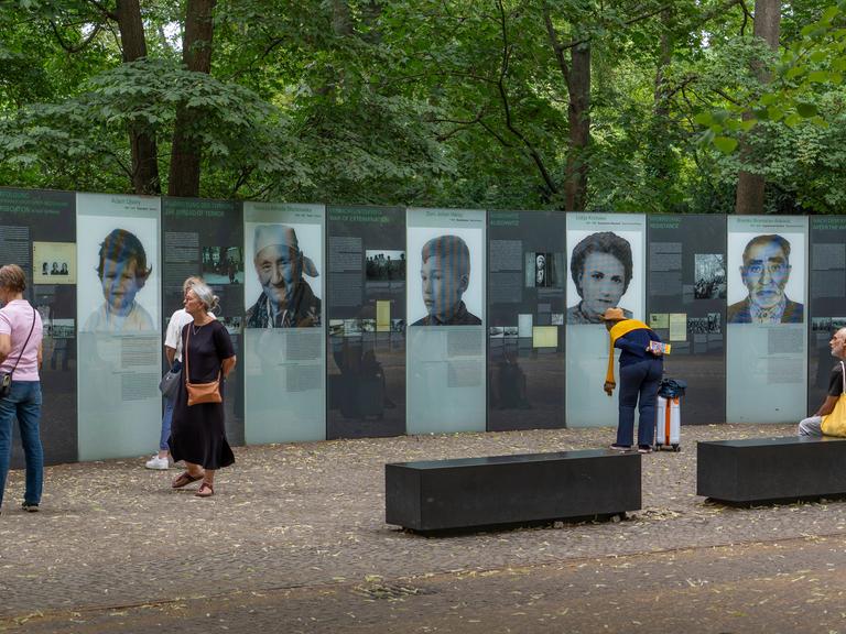 Mehrere Menschen stehen vor einem Denkmal, das an die von Nationalsozialisten ermordeten Sinti und Roma in Europa erinnert. Auf Gedenktafeln sind die Gesichter mehrerer Opfer und deren Geschichten abgebildet. 