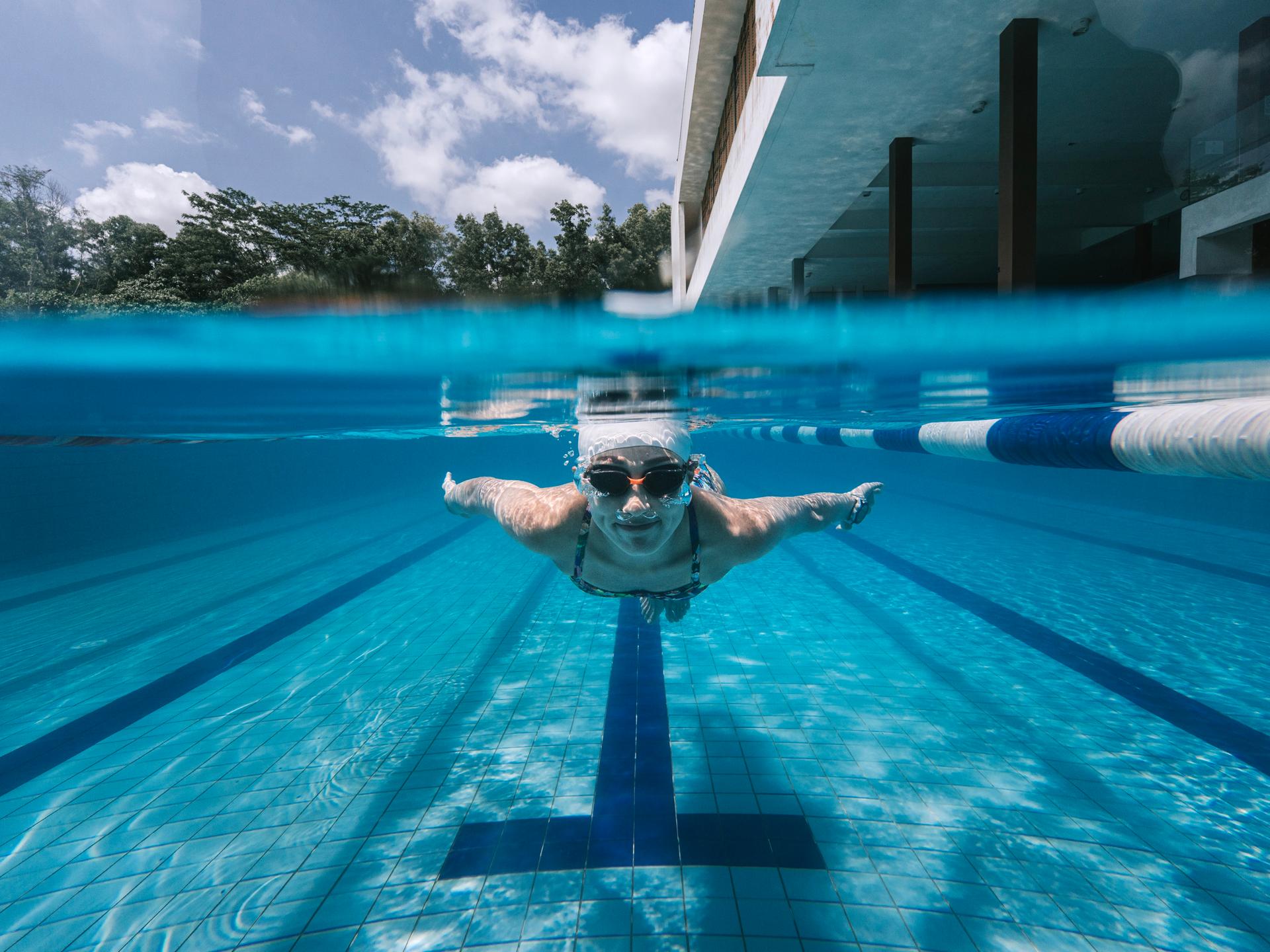 Eine Schwimmerin beim Kraulen im Schwimmbecken, vorn vorne aufgenommen, man sieht im split screen sowohl sie unter Wasser als auch den Hintergrund darüber