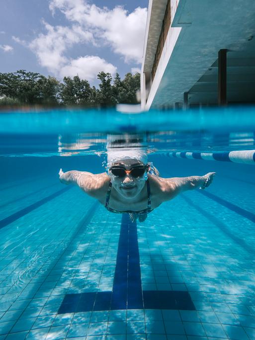 Eine Schwimmerin beim Kraulen im Schwimmbecken, vorn vorne aufgenommen, man sieht im split screen sowohl sie unter Wasser als auch den Hintergrund darüber