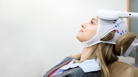 Eine Frau sitzt auf einem Behandlungsstuhl und schaut zur Decke. Auf ihrem Kopf trägt sie eine Bedeckung. Außerdem ist ein medizinisches Gerät auf ihren Kopf gerichtet.