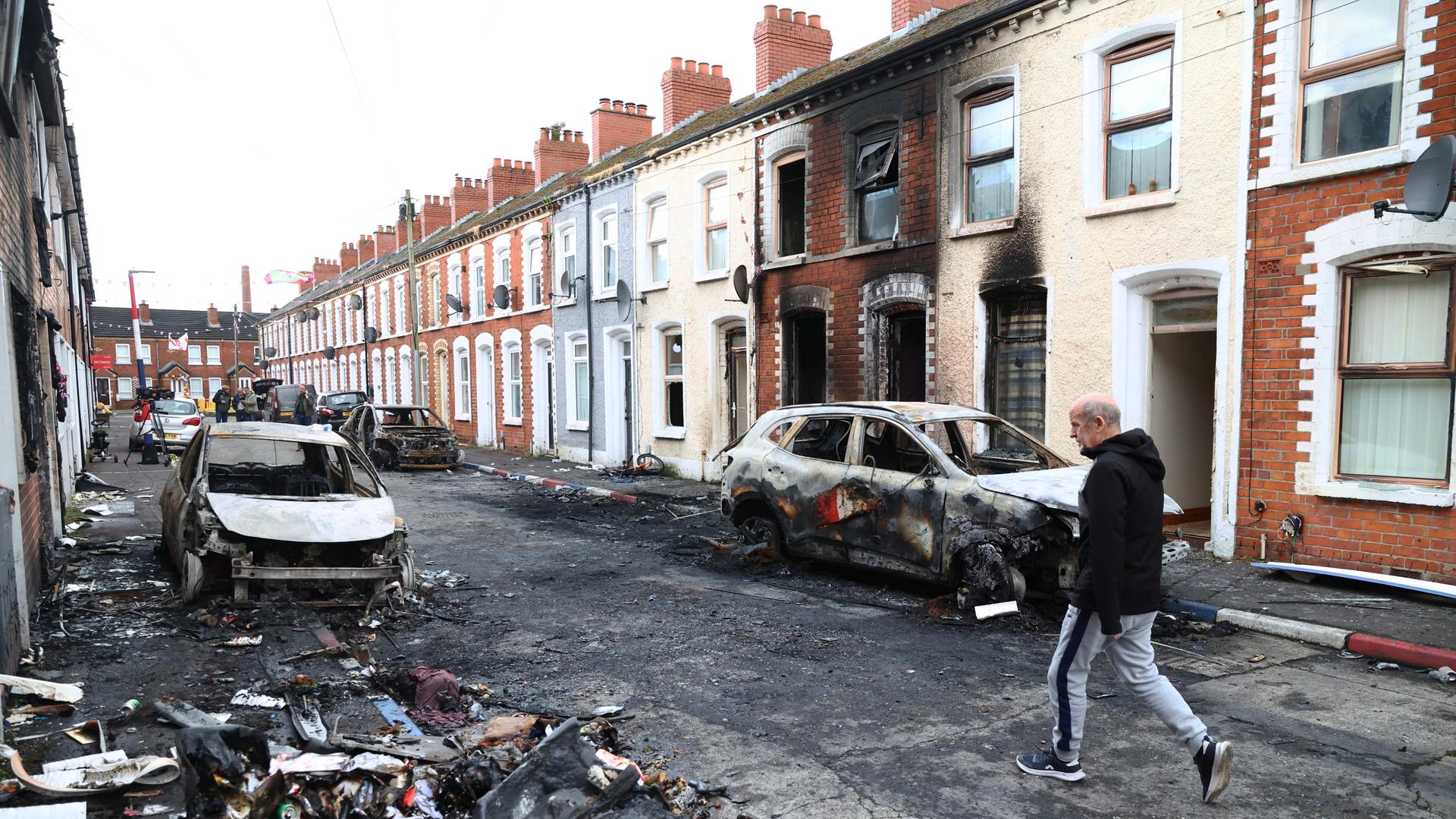 Ein Mann überquert in Belfast eine Straße. Dort stehen ausgebrannte PKW und Häuser. Ein Mann überquert in Belfast eine Straße. Dort stehen ausgebrannte PKW und Häuser.