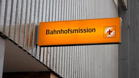 Beleuchtetes, oranges Schild der Münchner Bahnhofsmission vor einer grauen Wand. 