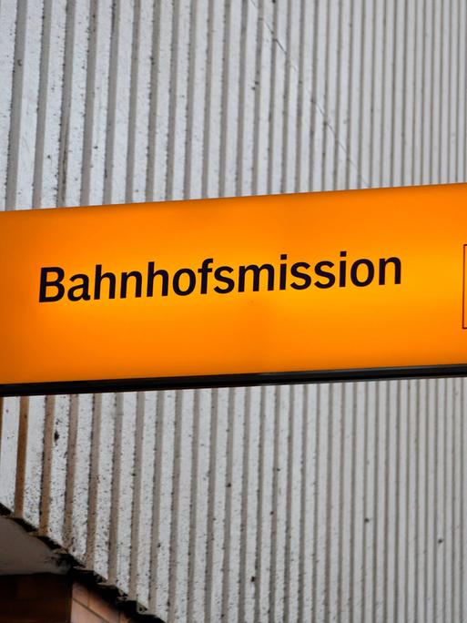 Beleuchtetes, oranges Schild der Münchner Bahnhofsmission vor einer grauen Wand. 