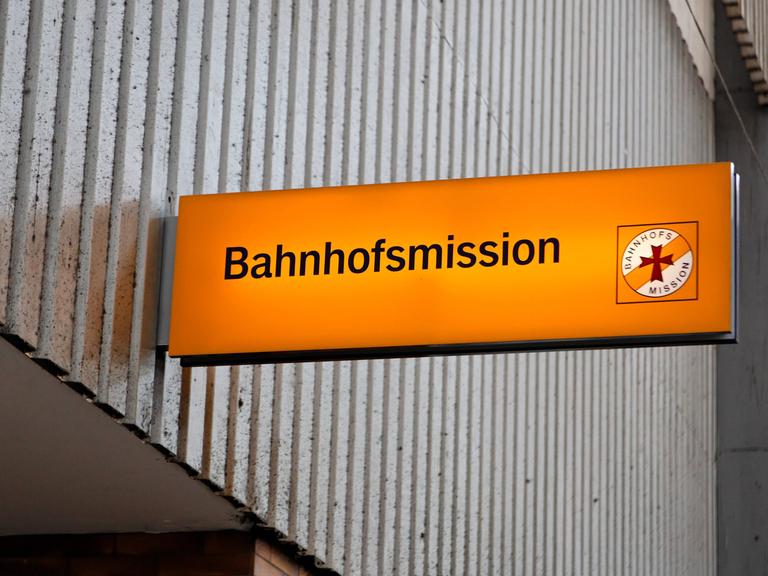 Beleuchtetes, oranges Schild der Münchner Bahnhofsmission vor einer grauen Wand. 