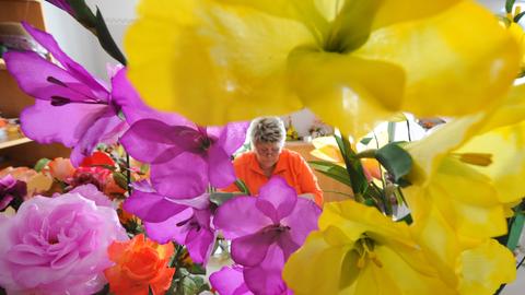 Im Vordergrund sind in der Schauwerkstatt "Deutsche Kunstblume Sebnitz" üppige, verschiedenfarbige Kunstblumen zu sehen, im Hintergrund eine Frau, die weitere Blumen fertigt.