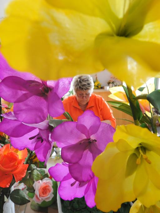 Im Vordergrund sind in der Schauwerkstatt "Deutsche Kunstblume Sebnitz" üppige, verschiedenfarbige Kunstblumen zu sehen, im Hintergrund eine Frau, die weitere Blumen fertigt.