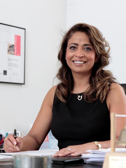 Nadia Qani sitzt am Schreibtisch in ihrem Büro und schaut freundlich in die Kamera.