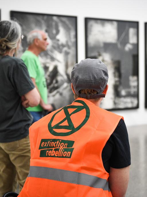 Führung im Musem Folkwang, im Vordergrund ist eine Person mit knalloranger "Extiction Rebellion"-Weste zu sehen, im Hintergrund eine Person, die ein Bild in der Ausstellung erläutert.