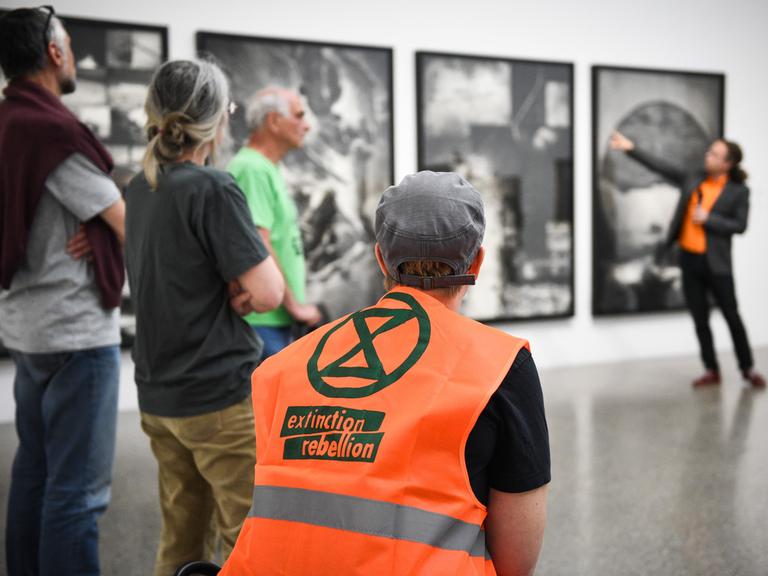 Führung im Musem Folkwang, im Vordergrund ist eine Person mit knalloranger "Extiction Rebellion"-Weste zu sehen, im Hintergrund eine Person, die ein Bild in der Ausstellung erläutert.