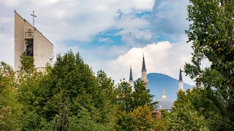 Im Stadtbild von Tirana sind über Bäumen der Turm einer Kirche und die Kuppel und Minarette einer Moschee zu sehen.
