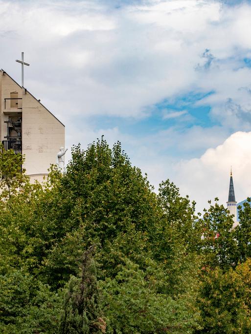 Im Stadtbild von Tirana sind über Bäumen der Turm einer Kirche und die Kuppel und Minarette einer Moschee zu sehen.