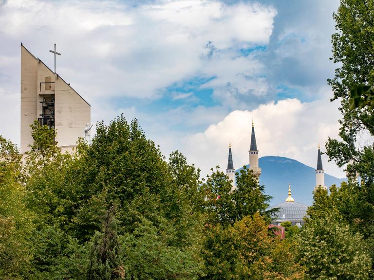 Im Stadtbild von Tirana sind über Bäumen der Turm einer Kirche und die Kuppel und Minarette einer Moschee zu sehen.