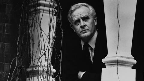 John le Carré im Jahr 2000 auf einem Schwarz-Weiß-Portrait in seinem Zuhause in Hampstead. Er schaut zwischen zwei Säulen hindurch. Er trägt einen dunklen Anzug und Krawatte und ein helles Hemd.
