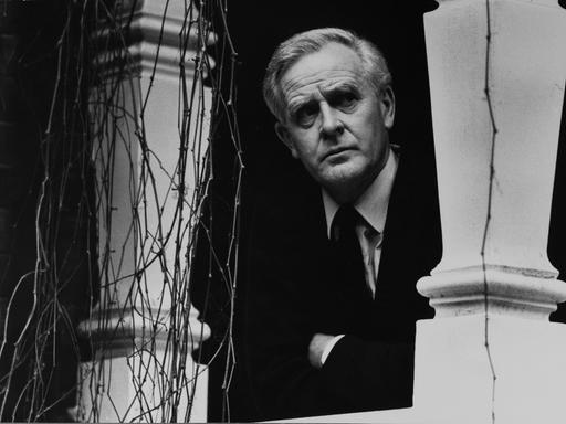 John le Carré im Jahr 2000 auf einem Schwarz-Weiß-Portrait in seinem Zuhause in Hampstead. Er schaut zwischen zwei Säulen hindurch. Er trägt einen dunklen Anzug und Krawatte und ein helles Hemd.