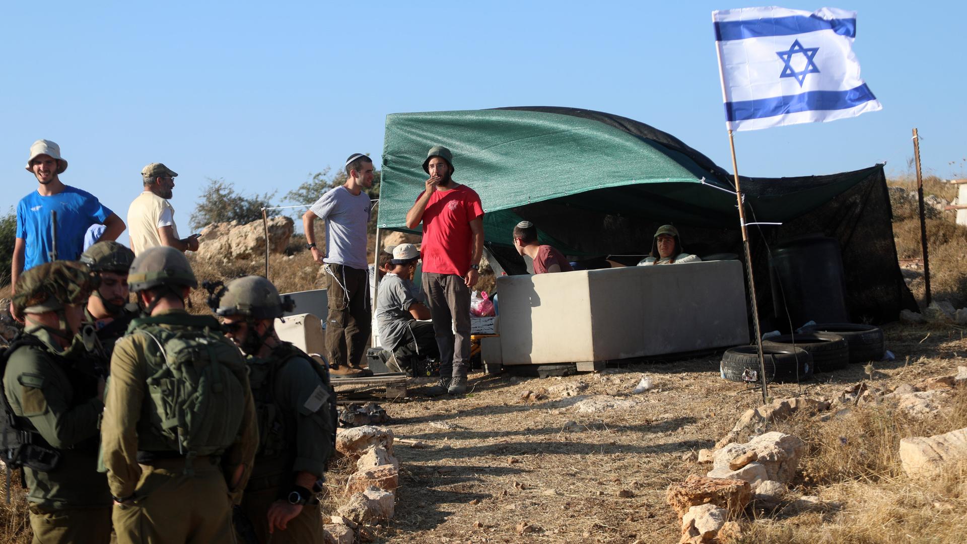 Über einem Zelt weht eine israelische Fahne; davor sind Siedler sowie israelische Soldaten zu sehen.
