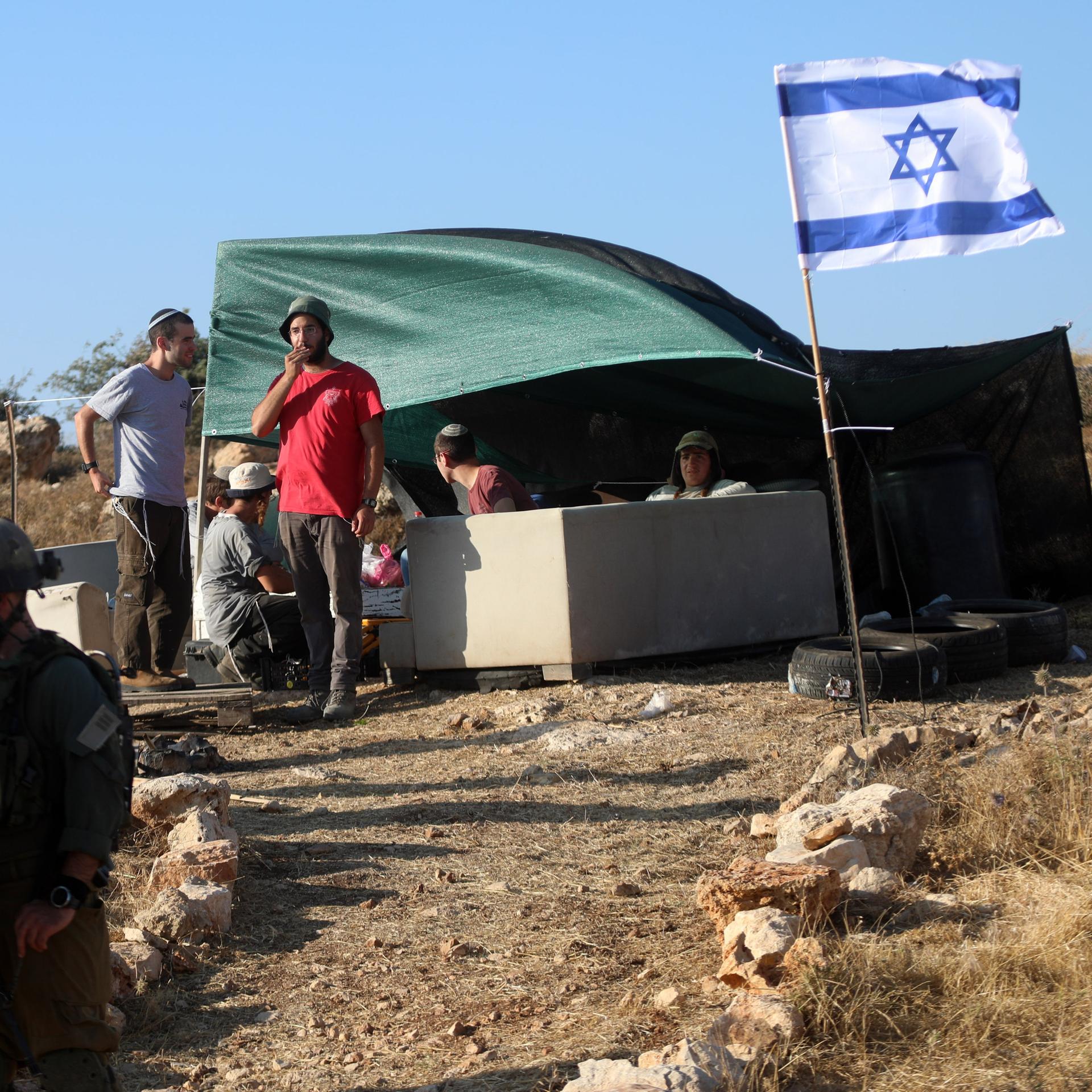 Über einem Zelt weht eine israelische Fahne; davor sind Siedler sowie israelische Soldaten zu sehen.