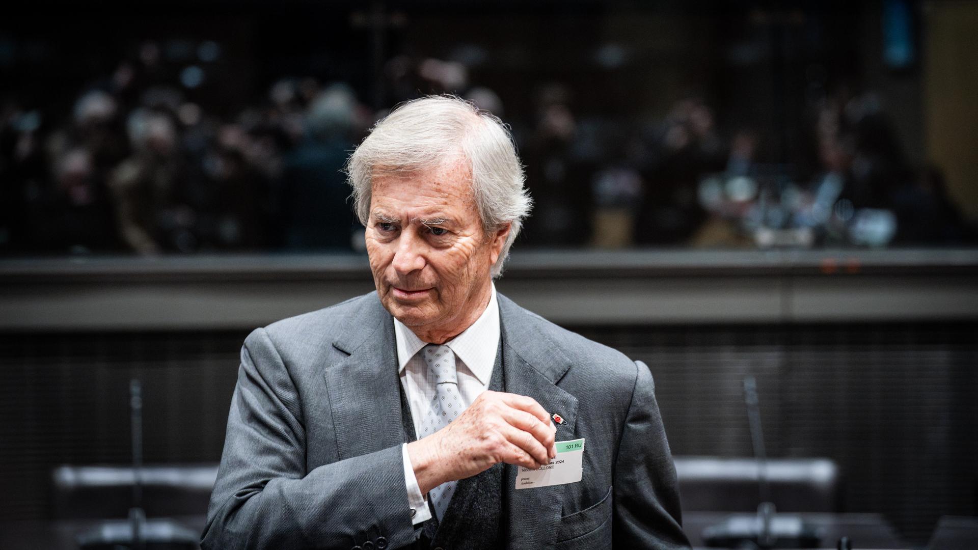 Vincent Bollore trägt einen grauen Anzug, weißes Hemd und eine helle gemusterte Krawatte. Er steht in einem Saal mit Tribüne im Hintergrund, auf der Menschen in der Unschärfe zu erkennen sind. Vincent Bollore trägt einen grauen Anzug, weißes Hemd und eine helle gemusterte Krawatte. Er steht in einem Saal mit Tribüne im Hintergrund, auf der Menschen in der Unschärfe zu erkennen sind.