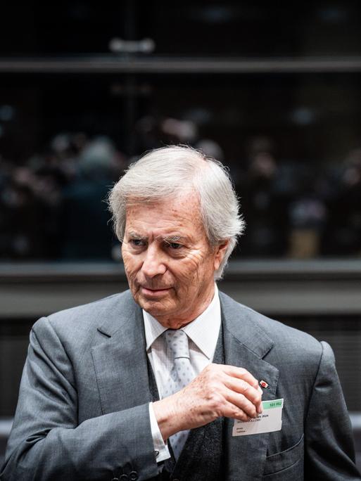 Vincent Bollore trägt einen grauen Anzug, weißes Hemd und eine helle gemusterte Krawatte. Er steht in einem Saal mit Tribüne im Hintergrund, auf der Menschen in der Unschärfe zu erkennen sind.