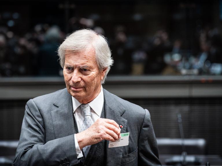 Vincent Bollore trägt einen grauen Anzug, weißes Hemd und eine helle gemusterte Krawatte. Er steht in einem Saal mit Tribüne im Hintergrund, auf der Menschen in der Unschärfe zu erkennen sind.