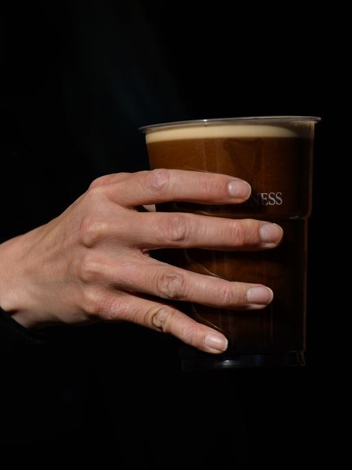 Eine Hand hält einen Plastikbecher mit dunklem Bier.