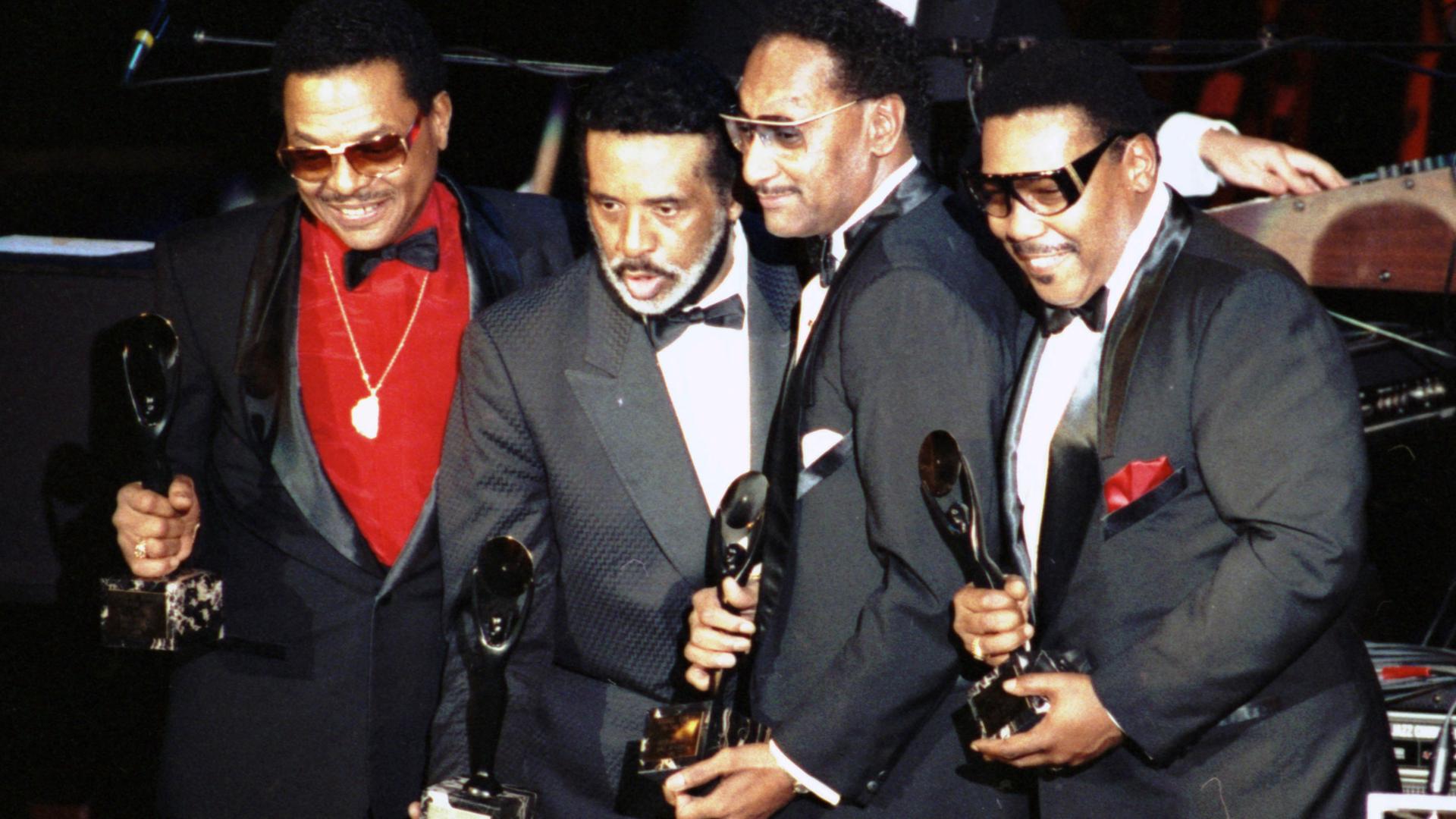 Das Bild zeigt die vier Mitglieder der Band Four Tops mit festlichen Anzügen und Auszeichnungen auf einer Bühne, sie freuen sich und lächeln Richtung Publikum.