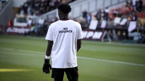 Ein Fußballer steht auf dem Feld und trägt ein weißes Trikot, auf dessen Rücken in schwarzen Buchstaben "No Violence" steht.