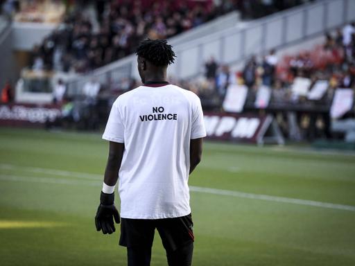 Ein Fußballer steht auf dem Feld und trägt ein weißes Trikot, auf dessen Rücken in schwarzen Buchstaben "No Violence" steht.