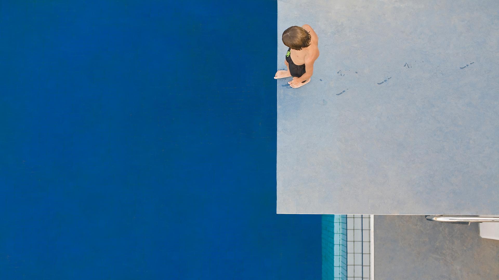 Ein Kind, das von oben zu sehen ist, steht auf einem Sprungturm und schaut auf das unter ihm liegende blaue Schwimmbecken. Ein Kind, das von oben zu sehen ist, steht auf einem Sprungturm und schaut auf das unter ihm liegende blaue Schwimmbecken.