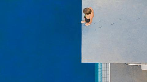 Ein Kind, das von oben zu sehen ist, steht auf einem Sprungturm und schaut auf das unter ihm liegende blaue Schwimmbecken.