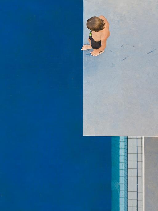 Ein Kind, das von oben zu sehen ist, steht auf einem Sprungturm und schaut auf das unter ihm liegende blaue Schwimmbecken.