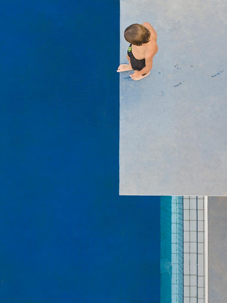 Ein Kind, das von oben zu sehen ist, steht auf einem Sprungturm und schaut auf das unter ihm liegende blaue Schwimmbecken.