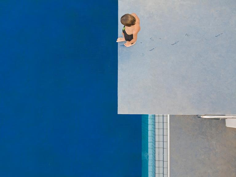 Ein Kind, das von oben zu sehen ist, steht auf einem Sprungturm und schaut auf das unter ihm liegende blaue Schwimmbecken.