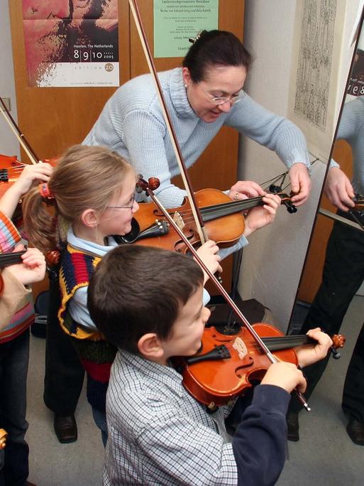 Geigenlehrerin erklärt am den kleinsten Geigenschülern vor dem Spiegel die richtige Haltung des Instruments. 