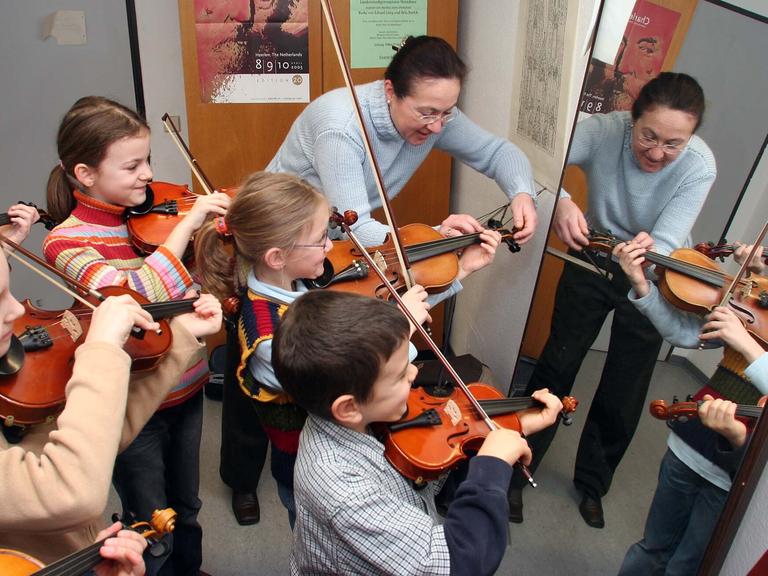 Geigenlehrerin erklärt am den kleinsten Geigenschülern vor dem Spiegel die richtige Haltung des Instruments. 