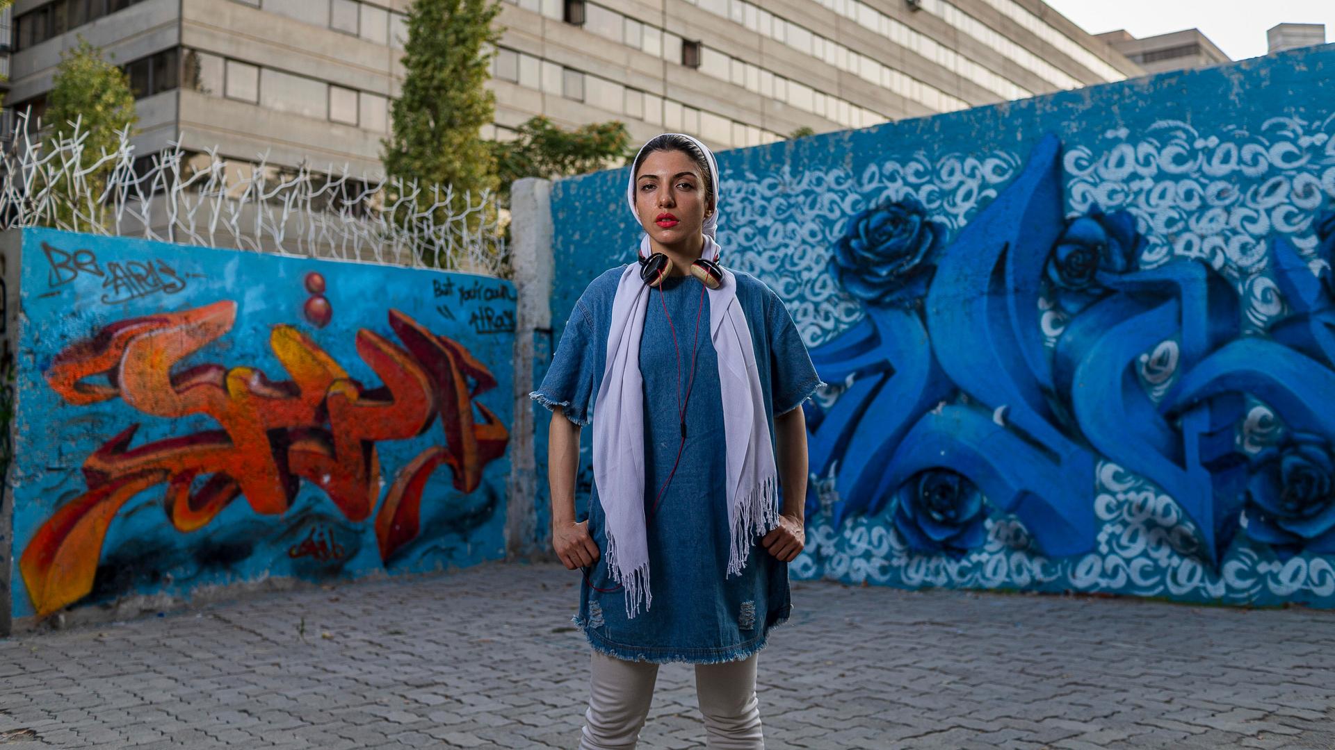 Die iranische Rapperin Justina steht vor einer mit Graffiti übersäten Hauswand im Teherander Stadtteil Ekbatan. Sie trägt ein blaues Jeanskleid, Kopfhörer und -tuch.