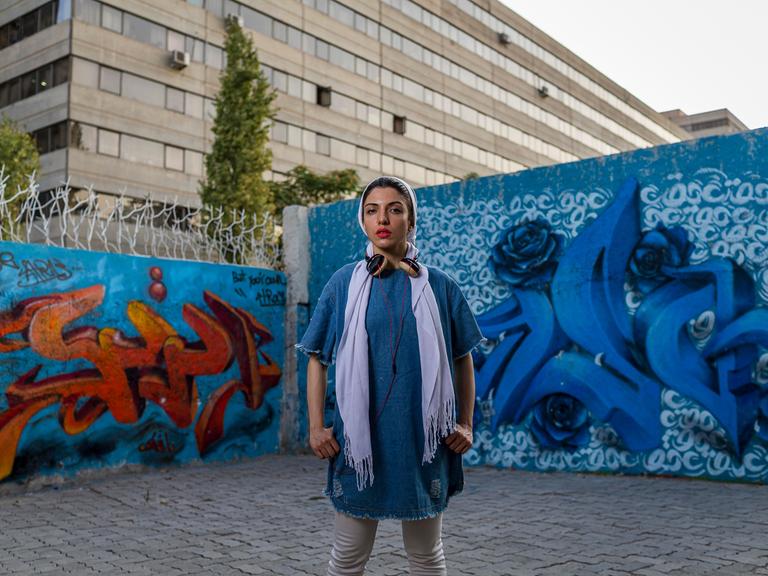 Die iranische Rapperin Justina steht vor einer mit Graffiti übersäten Hauswand im Teherander Stadtteil Ekbatan. Sie trägt ein blaues Jeanskleid, Kopfhörer und -tuch.